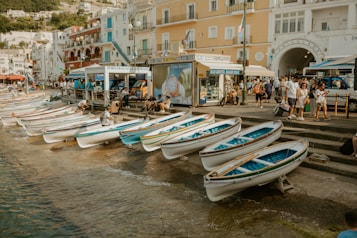 A seaside promenade features multiple small white and blue rowing boats lined up along the water's edge. People are seen walking and interacting near buildings that have a Mediterranean architectural style, painted in shades of white, yellow, and red. A sign advertising boat tours is visible next to a group of tourists, and the overall scene appears lively and bustling.