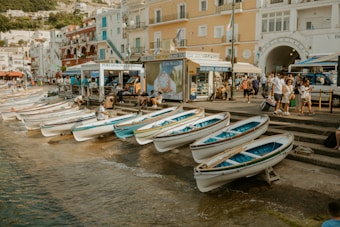 A seaside promenade features multiple small white and blue rowing boats lined up along the water's edge. People are seen walking and interacting near buildings that have a Mediterranean architectural style, painted in shades of white, yellow, and red. A sign advertising boat tours is visible next to a group of tourists, and the overall scene appears lively and bustling.