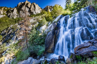 A scenic view of a waterfall cascading into a clear river surrounded by tall trees and rolling green hills under a bright sky.