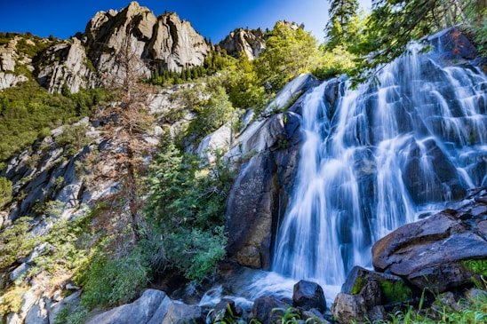 A scenic view of a waterfall cascading into a clear river surrounded by tall trees and rolling green hills under a bright sky.