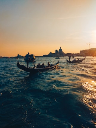 Gondolas glide along a shimmering body of water with a hazy silhouette of historic buildings under a warm sunset sky. The scene is tranquil with gentle waves reflecting the golden hues of the setting sun.