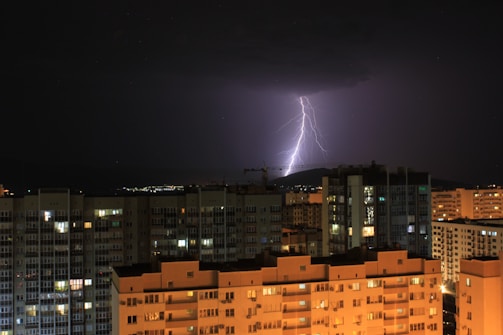 A nighttime urban scene with a dramatic lightning bolt illuminating the sky. The cityscape includes multiple high-rise apartment buildings with lights shining from windows, creating a contrast with the dark sky. The lightning is striking in the distance behind the buildings, casting an eerie glow over the area.