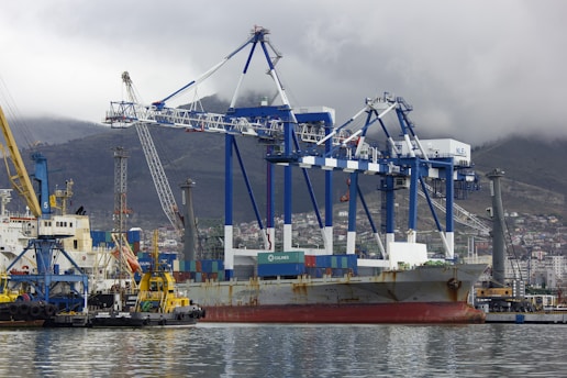 A large cargo ship docked at a port with several towering blue and white gantry cranes surrounding it. The ship is loaded with colorful shipping containers. In the foreground, a small yellow tugboat is assisting in docking. A hazy mountain range can be seen in the background under a cloudy sky.