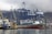 A large cargo ship docked at a port with several towering blue and white gantry cranes surrounding it. The ship is loaded with colorful shipping containers. In the foreground, a small yellow tugboat is assisting in docking. A hazy mountain range can be seen in the background under a cloudy sky.