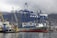 A large cargo ship docked at a port with several towering blue and white gantry cranes surrounding it. The ship is loaded with colorful shipping containers. In the foreground, a small yellow tugboat is assisting in docking. A hazy mountain range can be seen in the background under a cloudy sky.