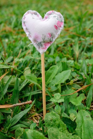 A small heart-shaped fabric decoration on a stick placed in lush green grass. The fabric features a soft floral pattern with pink and white colors, giving it a gentle, handcrafted appearance.