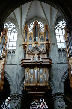 A grand and ornate pipe organ is mounted on a stone wall within a gothic cathedral, showcasing intricate craftsmanship with numerous pipes and wooden structures. The surrounding architecture features detailed stone carvings, large arched windows with delicate tracery, and stained glass elements that contribute to the majestic atmosphere of the setting.