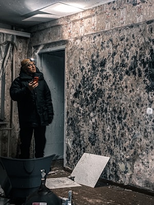 A man stands inside a room with peeling, mold-covered wallpaper, looking at a smartphone. He is wearing a winter coat and a headlamp, indicating poor lighting or working conditions. The floor is cluttered with debris and broken tile pieces.
