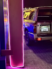 A parked Nissan Patrol vehicle with a visible license plate. The scene features vibrant neon purple and pink lighting, which casts reflections on the car. The ground is paved with bricks, and string lights can be seen in the background.