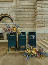 Several mailboxes stand against a textured stone wall, accompanied by vibrant floral arrangements. The flowers are predominantly blue, yellow, and red, spilling gracefully from the mailboxes and onto the ground.