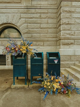Several mailboxes stand against a textured stone wall, accompanied by vibrant floral arrangements. The flowers are predominantly blue, yellow, and red, spilling gracefully from the mailboxes and onto the ground.