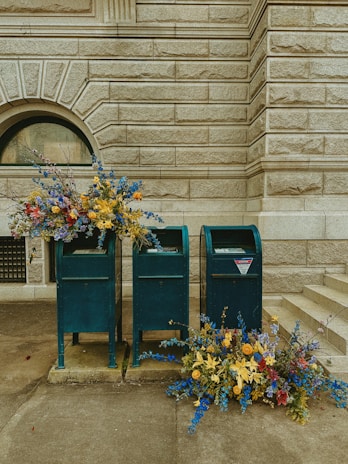 Several mailboxes stand against a textured stone wall, accompanied by vibrant floral arrangements. The flowers are predominantly blue, yellow, and red, spilling gracefully from the mailboxes and onto the ground.