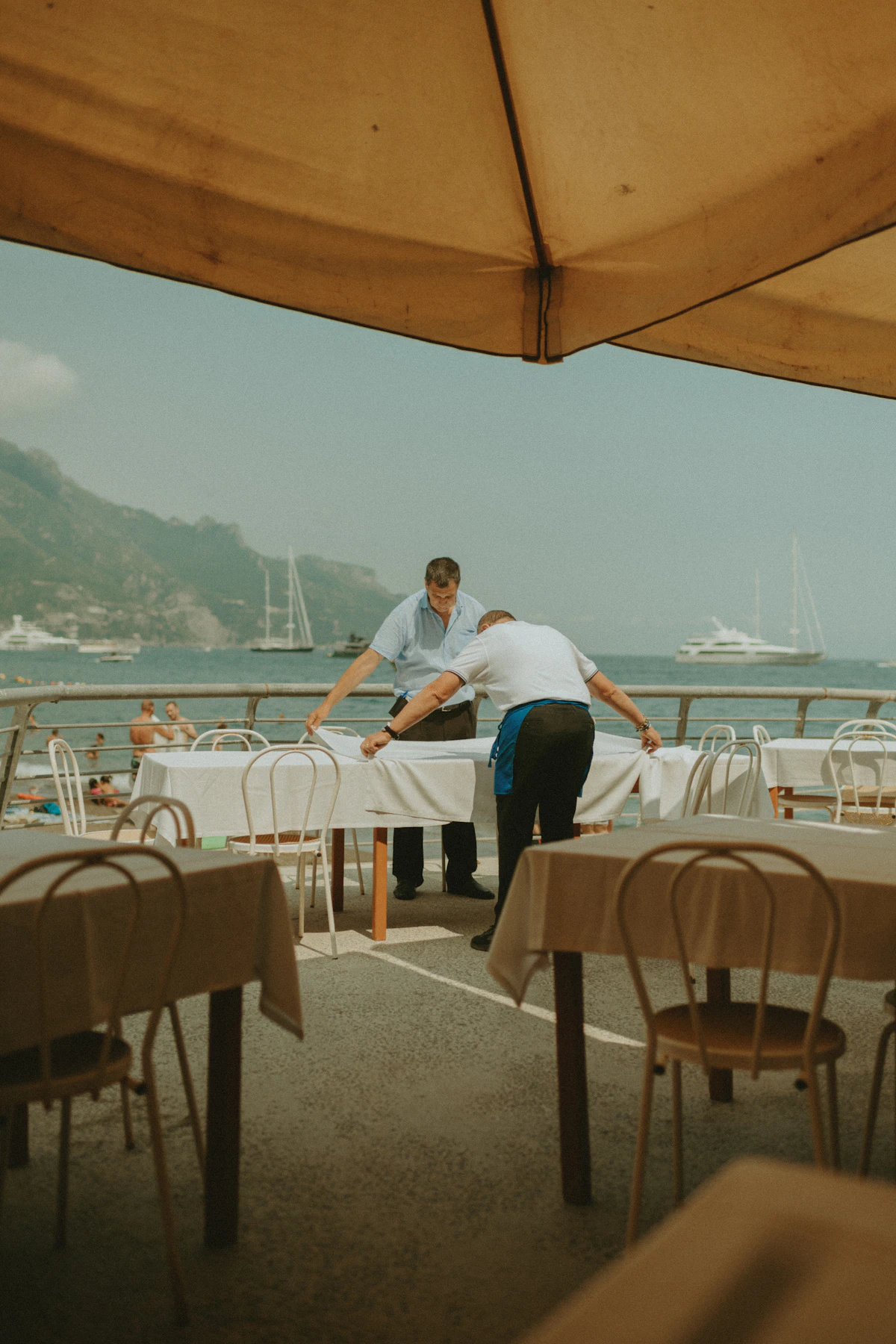 Two staff reset white-clothed tables on an empty seaside terrace under an awning, boats on the water behind them.