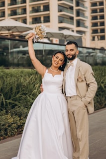 A joyful couple in wedding attire poses outdoors. The woman is wearing a white wedding dress and holding a bouquet, while the man is dressed in a beige suit with a white shirt and bow tie. They are standing close together with greenery and a modern building in the background.