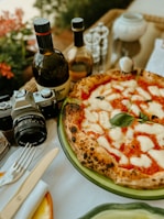 A delicious pizza topped with melted mozzarella and basil sits on a green plate alongside a vintage camera, two bottles of olive oil or wine, and some tableware including a knife and fork. The background features some red flowers and blurred elements, suggesting an outdoor dining setting.