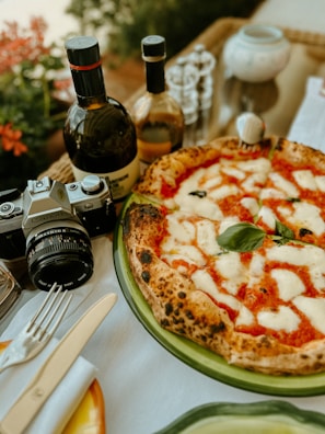 A delicious pizza topped with melted mozzarella and basil sits on a green plate alongside a vintage camera, two bottles of olive oil or wine, and some tableware including a knife and fork. The background features some red flowers and blurred elements, suggesting an outdoor dining setting.