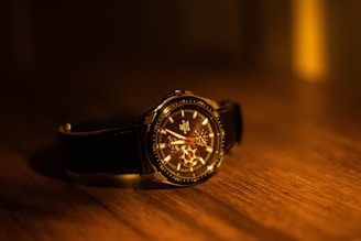 A luxury wristwatch with a leather band resting on a wooden surface. The watch face is intricately detailed with visible gears and hands, illuminated by warm, ambient lighting that casts soft shadows.
