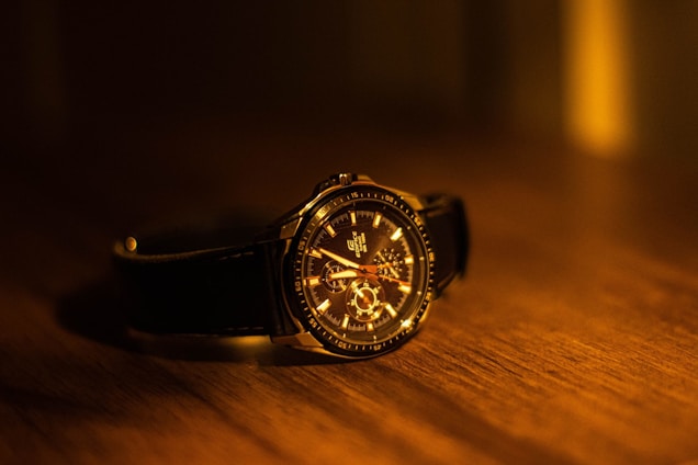 A luxury wristwatch with a leather band resting on a wooden surface. The watch face is intricately detailed with visible gears and hands, illuminated by warm, ambient lighting that casts soft shadows.