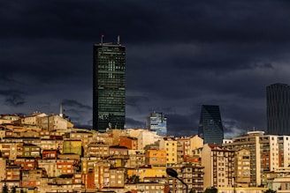 A dense cluster of residential buildings with a variety of colors and architectural styles stands in the foreground. The backdrop features several modern skyscrapers, including a tall, dark tower. The sky above is overcast, casting a dramatic shadow over the cityscape.