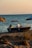 A small motorboat is parked on a sandy beach with gentle waves in the background. The boat has a dark hull and is equipped with life rings and a small outboard motor. There are beach umbrellas partially visible, and sparse grass grows in the sand nearby, creating a serene sunset scene.