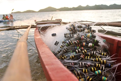 A fishing boat filled with tangled fishing nets and colorful floaters sits by the shore. The background shows a calm sea and distant hills under a soft-lit sky. A small group of people can be seen kayaking in the distance.