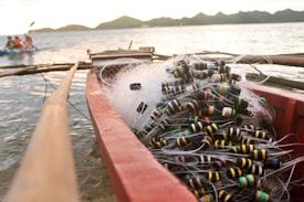 A fishing boat filled with tangled fishing nets and colorful floaters sits by the shore. The background shows a calm sea and distant hills under a soft-lit sky. A small group of people can be seen kayaking in the distance.