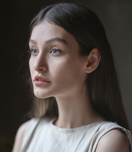 A portrait of a young woman with long dark hair wearing a light-colored sleeveless top. She has an expression of calm contemplation, with soft lighting highlighting her features against a blurred, dark background.
