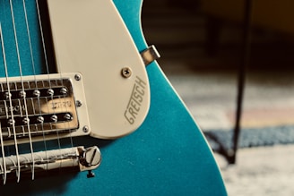 Close-up view of an electric guitar featuring a portion of the body and strings. The guitar has a shiny teal finish with chrome hardware. A white pickguard is visible with the brand name engraved on it.