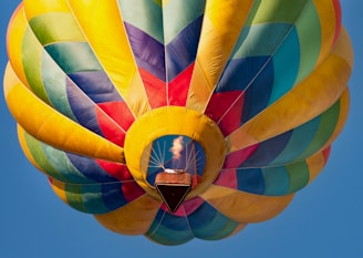 A vibrant hot air balloon with a patchwork design of colorful panels including blue, yellow, green, and red is floating against a clear blue sky. The balloon’s basket is visible with a flame emitting heat to lift the balloon.