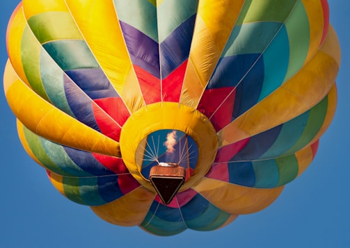A vibrant hot air balloon with a patchwork design of colorful panels including blue, yellow, green, and red is floating against a clear blue sky. The balloon’s basket is visible with a flame emitting heat to lift the balloon.