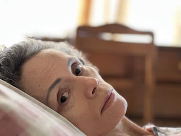 A person with short, curly gray hair rests their head on a pillow indoors. The background is softly blurred, with an emphasis on wooden furniture and warm lighting.