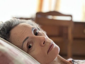 A person with short, curly gray hair rests their head on a pillow indoors. The background is softly blurred, with an emphasis on wooden furniture and warm lighting.