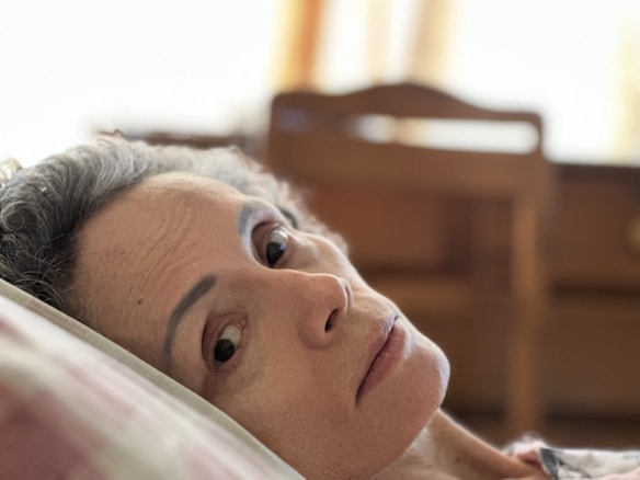 A person with short, curly gray hair rests their head on a pillow indoors. The background is softly blurred, with an emphasis on wooden furniture and warm lighting.