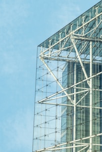 A modern architectural structure features a glass facade with visible steel beams and supports, offering a transparent view of the building's interior framework against a clear blue sky. The design emphasizes sleek lines and industrial elements.