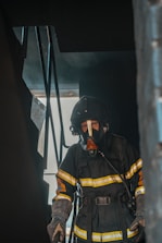 A firefighter in full protective gear, including a mask and helmet, stands inside a dark, smoke-filled environment near a staircase. Light filters in from a doorway or window, casting shadows on the scene.