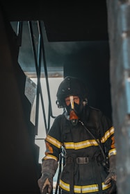 A firefighter in full protective gear, including a mask and helmet, stands inside a dark, smoke-filled environment near a staircase. Light filters in from a doorway or window, casting shadows on the scene.