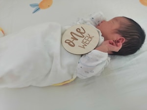 A newborn baby wrapped in a white blanket is lying on a bed. The baby has a wooden plaque resting on them with the words 'one week' engraved on it. The baby is wearing a white onesie with small patterns on it and appears to be sleeping peacefully. The bedding features soft colors and a cute design, adding to the serene setting.