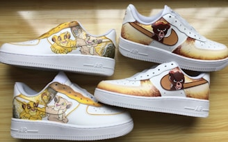 Four white sneakers feature colorful cartoon designs along the sides. The designs include animated characters in shades of yellow, brown, and cream, set against a vibrant and playful background. The sneakers are arranged on a light wood surface, showcasing both side and top views.
