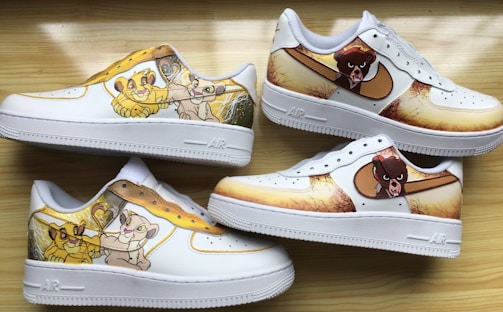 Four white sneakers feature colorful cartoon designs along the sides. The designs include animated characters in shades of yellow, brown, and cream, set against a vibrant and playful background. The sneakers are arranged on a light wood surface, showcasing both side and top views.