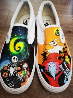 A pair of white slip-on shoes featuring colorful, hand-painted designs inspired by a Halloween-themed animated film. The left shoe displays a spooky night scene with swirling colors, featuring characters with distinctive skeleton and rag doll appearances surrounded by pumpkins. The right shoe shows a vibrant Halloween scene with a ghost dog and characters with exaggerated features and vivid costumes.