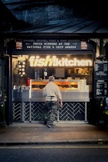 A person stands in front of a fish and chips takeaway shop with a brightly lit sign reading 'fish! kitchen'. The shop exterior features metal and brick accents, with awards displayed above the entrance, indicating recognition for their fish and chips. The scene is set during the evening, as the street in front is dimly lit.