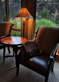 Two upholstered chairs are placed around a wooden table near a window with a view of outdoor greenery. A classic-style lamp with a pinecone accent casts a warm glow, and a dark leather bag is resting on one of the chairs.