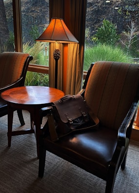 Two upholstered chairs are placed around a wooden table near a window with a view of outdoor greenery. A classic-style lamp with a pinecone accent casts a warm glow, and a dark leather bag is resting on one of the chairs.