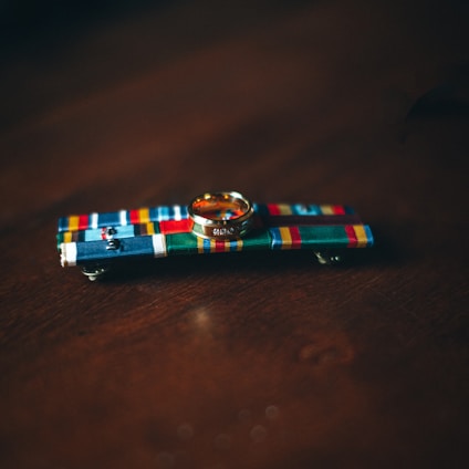 A gold ring rests on a colorful military ribbon bar with various patterns and stripes against a dark wooden surface.