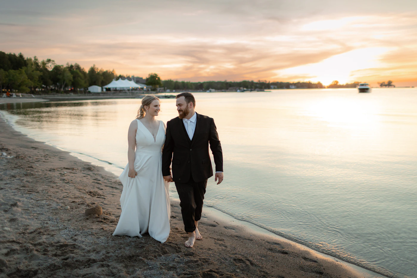 Bride in white wedding gown and groom walking along ocean beach shoreline at sunset with dramatic golden sky behind them