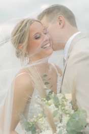 A joyful couple is surrounded by a soft veil, with the woman smiling radiantly and holding a bouquet of flowers. The man leans in closely, suggesting intimacy and affection.