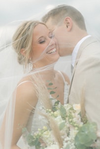 A joyful couple is surrounded by a soft veil, with the woman smiling radiantly and holding a bouquet of flowers. The man leans in closely, suggesting intimacy and affection.