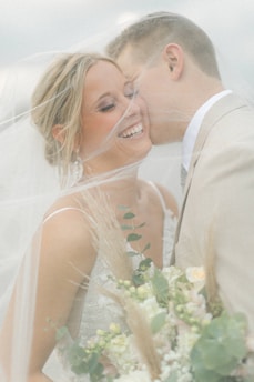 A joyful couple is surrounded by a soft veil, with the woman smiling radiantly and holding a bouquet of flowers. The man leans in closely, suggesting intimacy and affection.