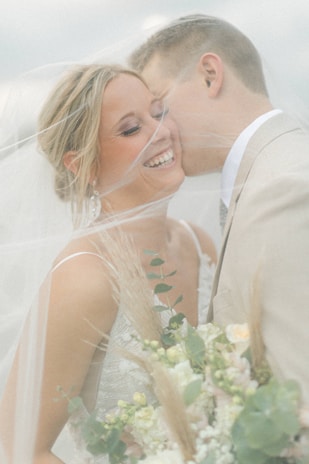 A joyful couple is surrounded by a soft veil, with the woman smiling radiantly and holding a bouquet of flowers. The man leans in closely, suggesting intimacy and affection.