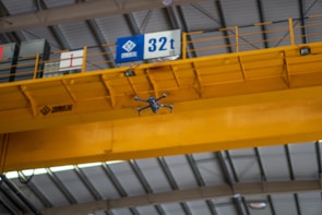 A drone is flying indoors near a large yellow overhead crane with a visible signage indicating 32t. The ceiling structure features metal beams and industrial lighting.